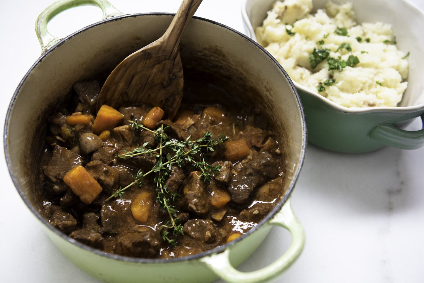 Beef stew with carrots and herbs in a green pot with mashed potatoes.