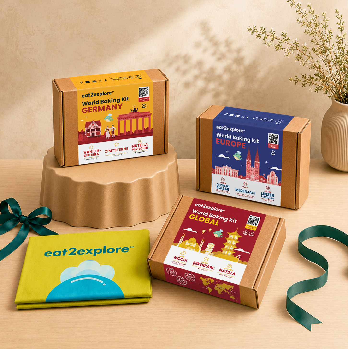Three eat2explore World Baking Kits in different regions (Germany, Europe, and Global) are displayed on a wooden table with a beige pedestal, alongside a green ribbon and a folded green apron on a light wood background.