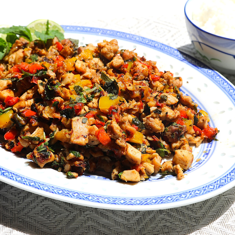 A close-up of a prepared food dish on a white plate with blue trim, showing cooked chicken pieces mixed with chopped vegetables including red, yellow, and green peppers and herbs. A lime wedge and a small bowl of white rice are partially visible in the background.