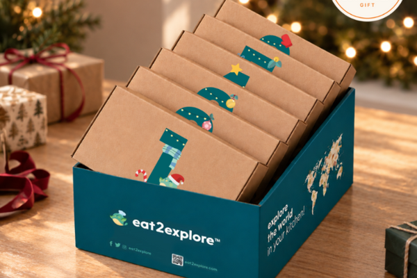 A teal and kraft cardboard gift box filled with multiple small brown snack boxes, displayed on a wooden table with holiday decorations in the background. The front box shows the Eat2Explore logo and name.