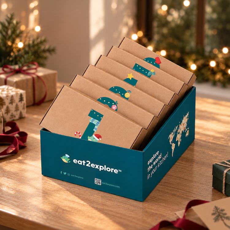 A teal Eat2explore gift box filled with multiple brown kraft mailer envelopes decorated with Christmas tree illustrations, displayed on a wooden table with holiday decorations in the background.