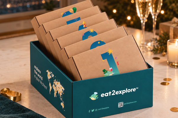 A teal and kraft paper gift box labeled eat2explore, filled with numbered travel-sized snack packs, set on a decorated holiday table with champagne flutes, candles, and gold coins in the background.