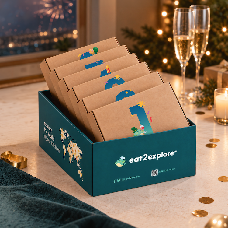 A teal and kraft paper gift box labeled eat2explore, filled with numbered travel-sized snack packs, set on a decorated holiday table with champagne flutes, candles, and gold coins in the background.