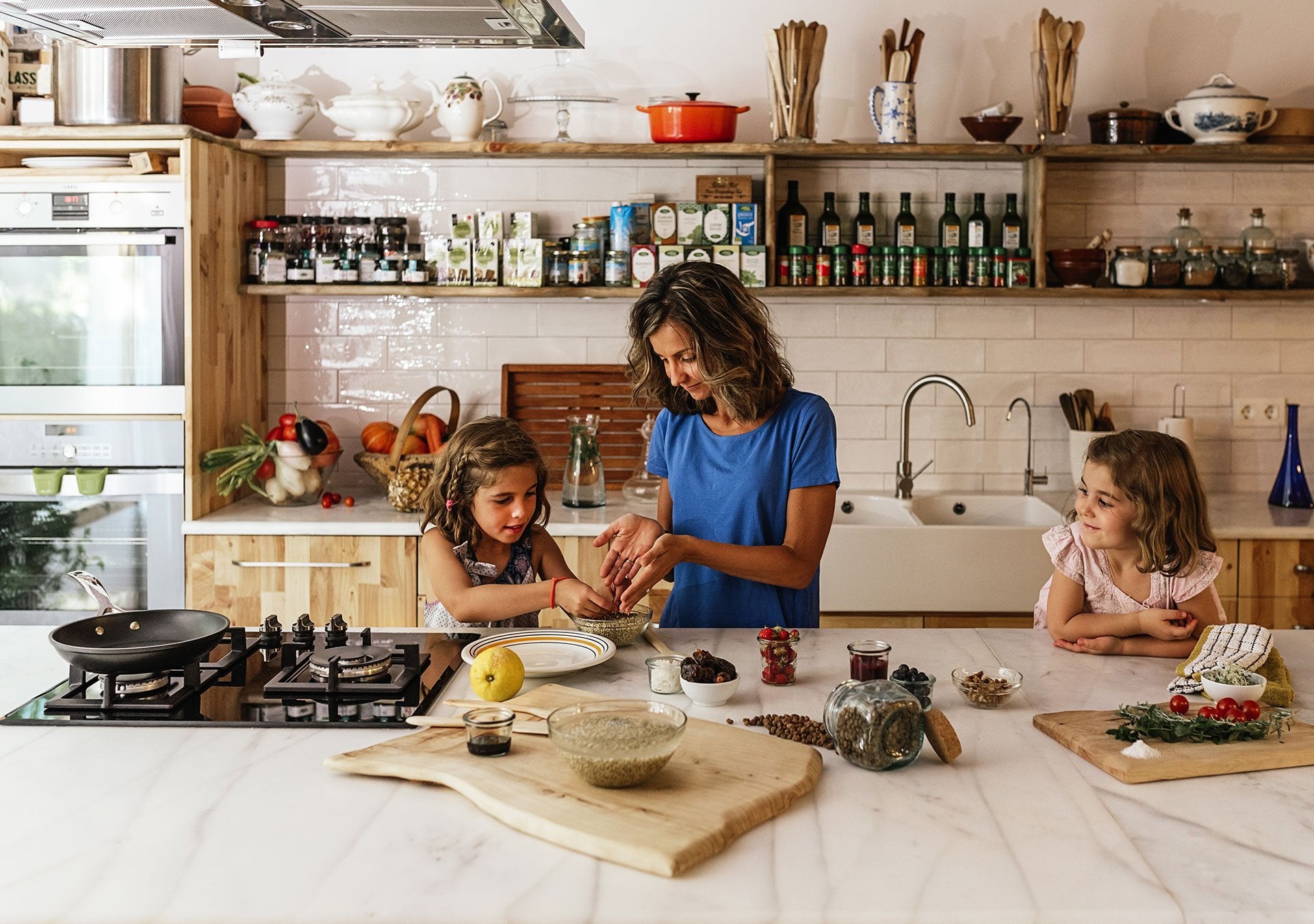 A mother and two daughters cooking together in a well-organized kitchen with various ingredients and cooking utensils on the counter and shelves.