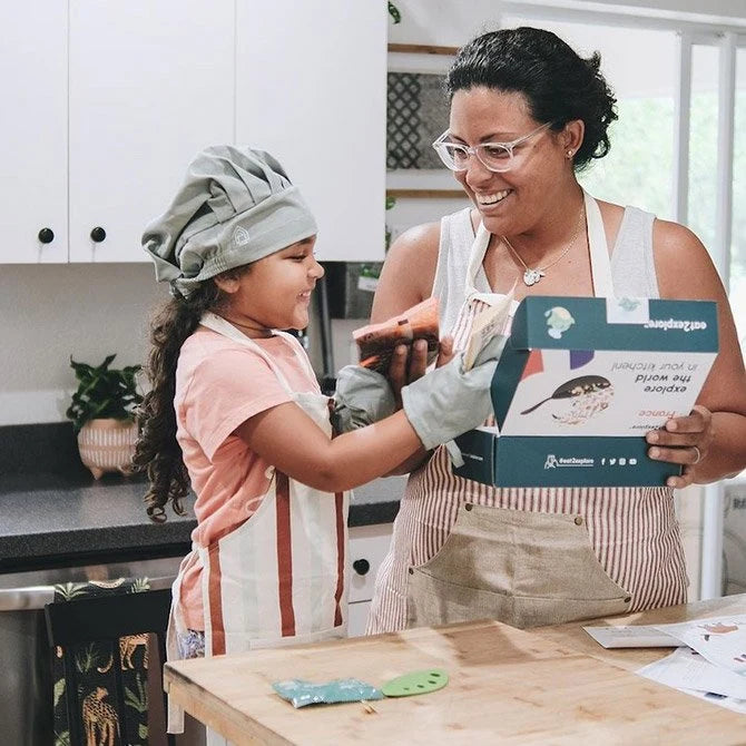 A mother and daughter smiling while cooking together in the kitchen, with the daughter wearing a chef's hat and apron and the mother holding a cooking kit box.